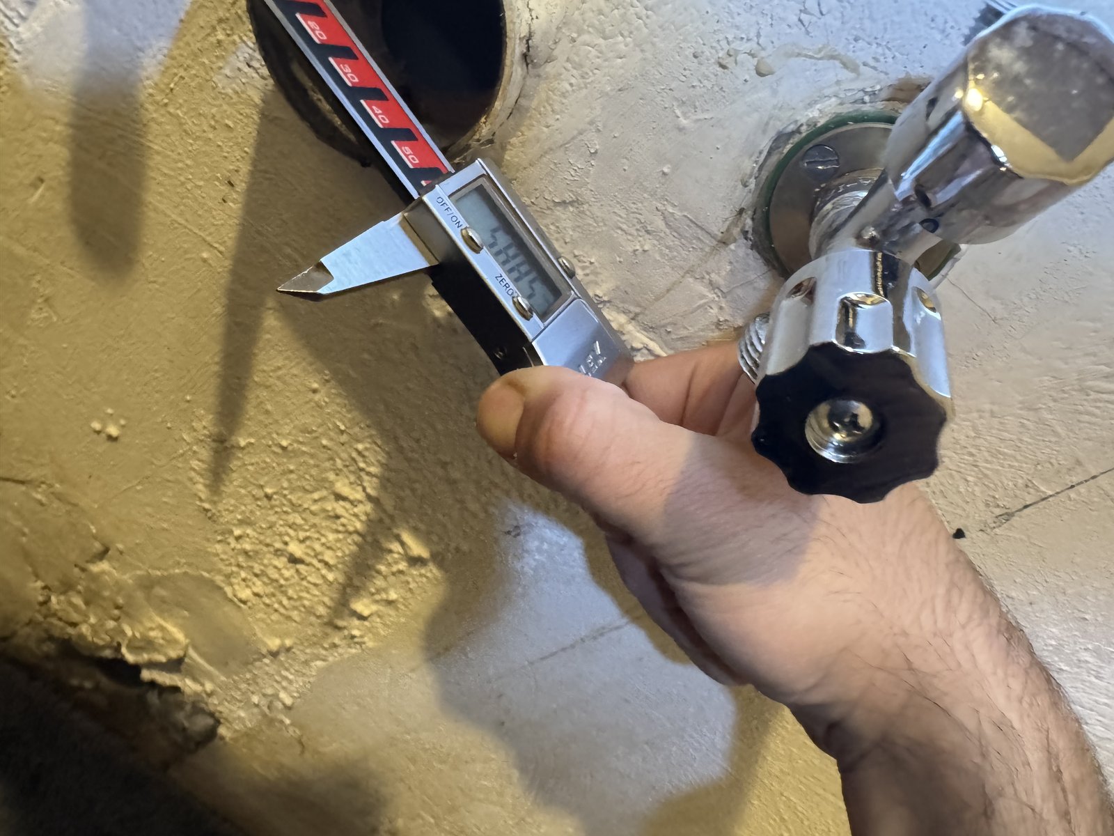 A hand holding a digital caliper to measure a pipe opening next to a wall valve