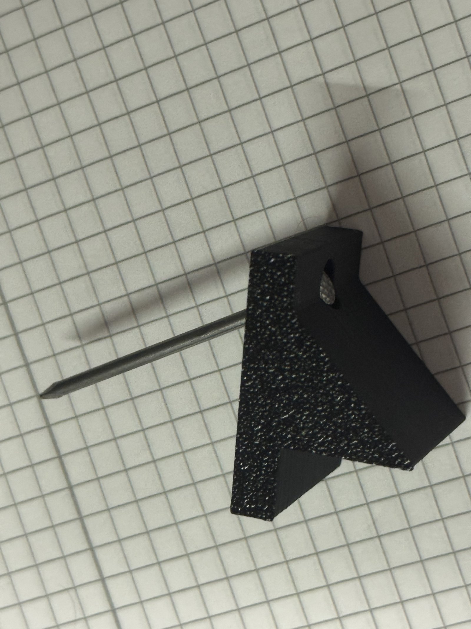A black 3D printed angle bracket with a textured surface lying next to a nail on a grid mat