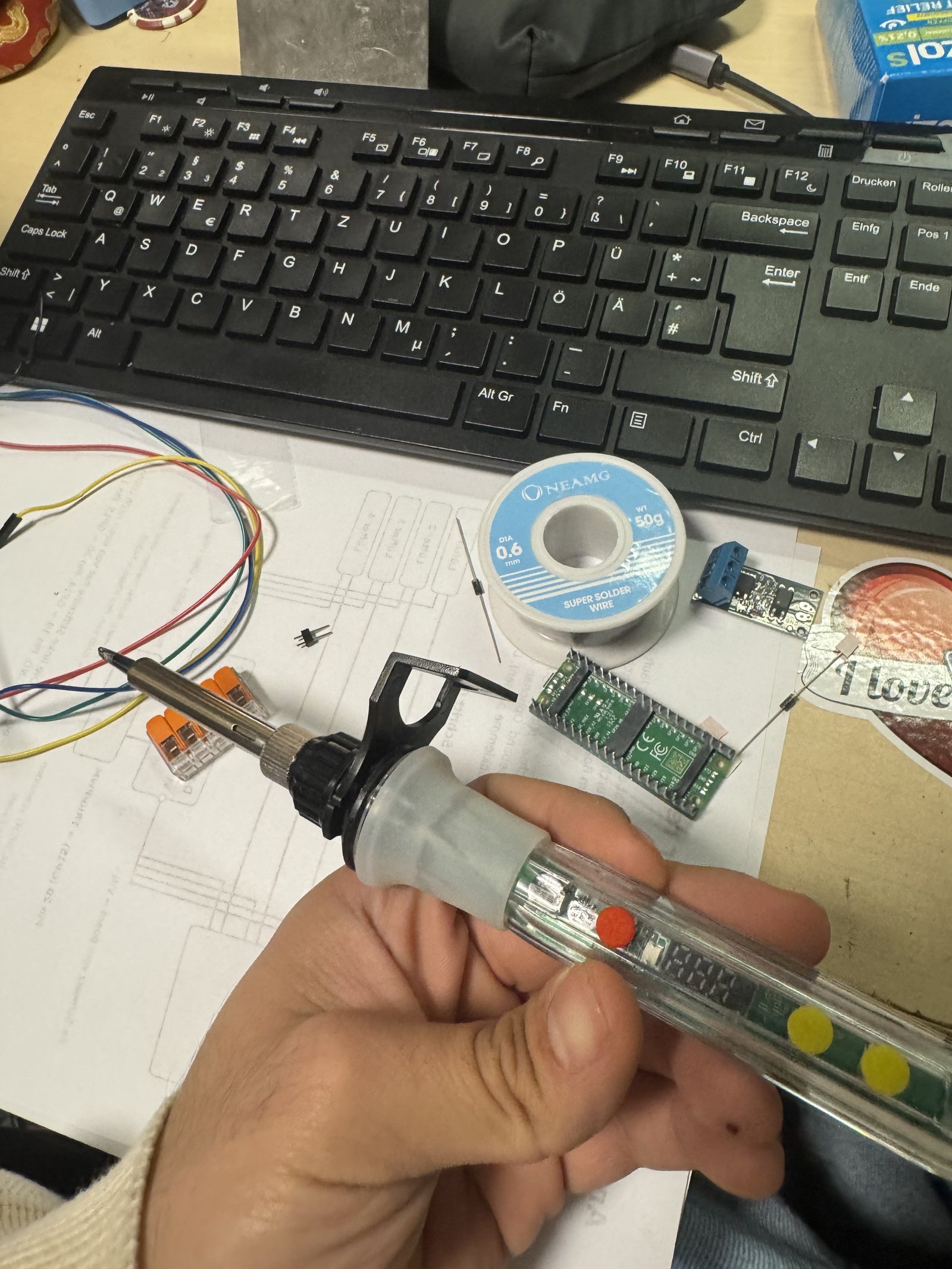 A hand holding a soldering iron above a desk with solder wire, a keyboard and small circuit boards